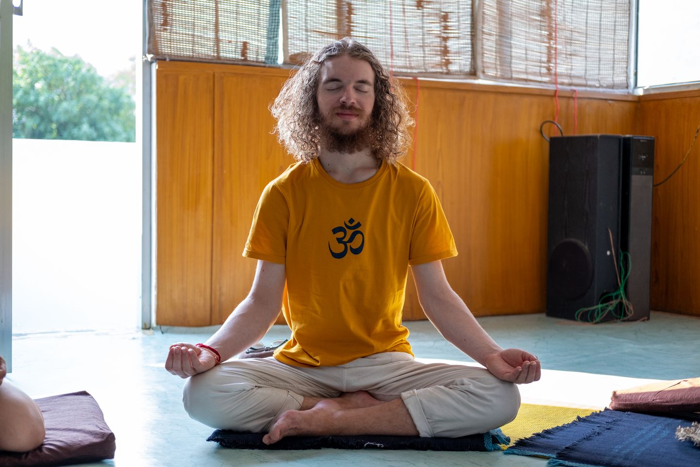 Relaxed young man meditating in a serene indoor yoga setting, wearing a yellow shirt with an Om symbol.